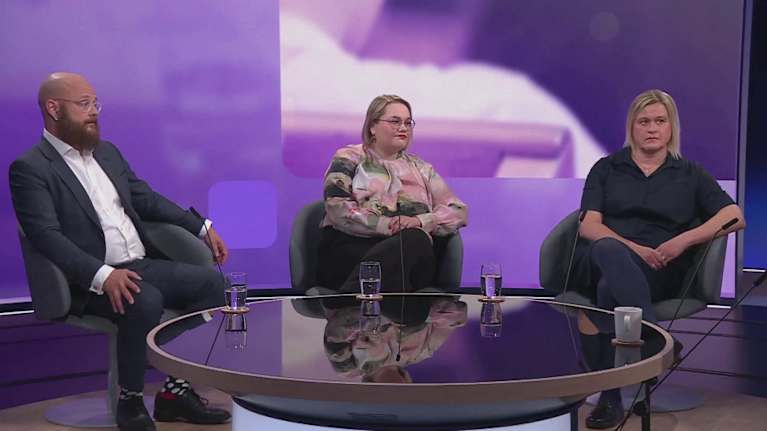 Two women and a man sit at a low coffee table in a television studio with a purple background.