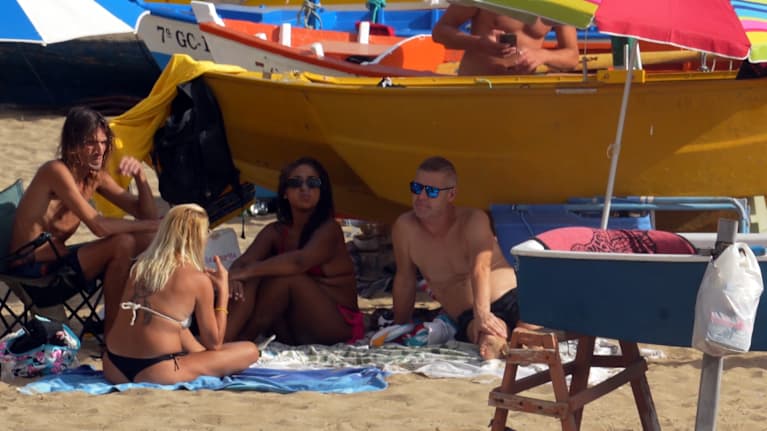 Cuatro jóvenes adultos en traje de baño se sientan en la playa bajo una sombrilla.