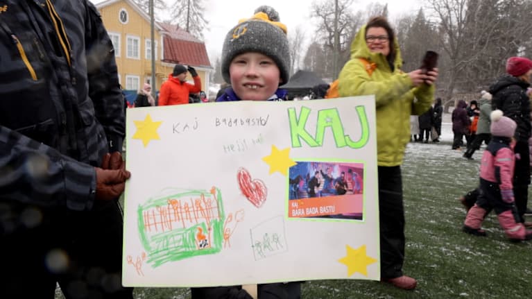 A young boy in a grey knit cap holding up a hand-drawn sign honouring KAJ.