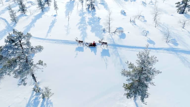 Ilmakuvassa joulupukki matkustaa kahden poron kanssa lumisessa maisemassa.