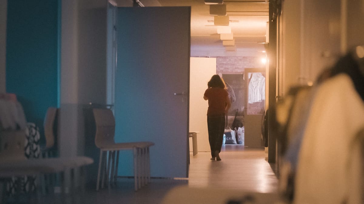 Asuntolan hämärä käytävä, jossa huoneiden ovet ovat auki. Yksi ihminen kävelee käytävän päässä.