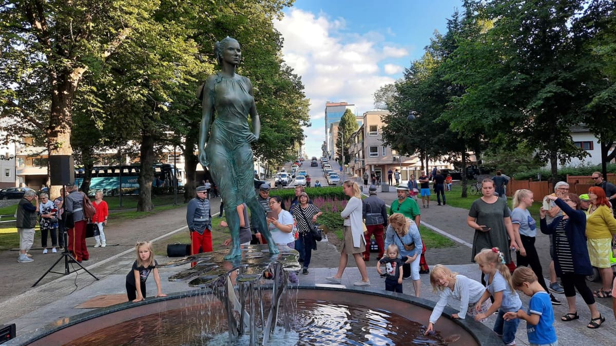 Naista esittävä tummahko patsas. Ihmisiä ympärillä kesäisessä maisemassa.