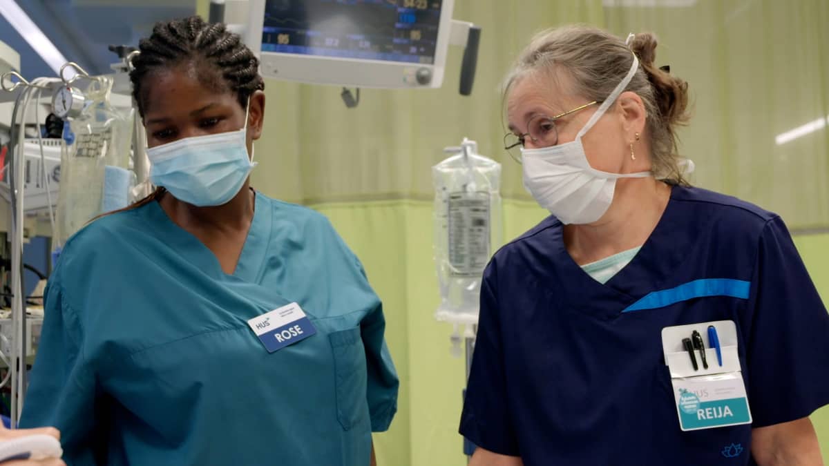 Two nurses of different ages and ethnic background wearing face masks and blue shirts standing in a hospital room.