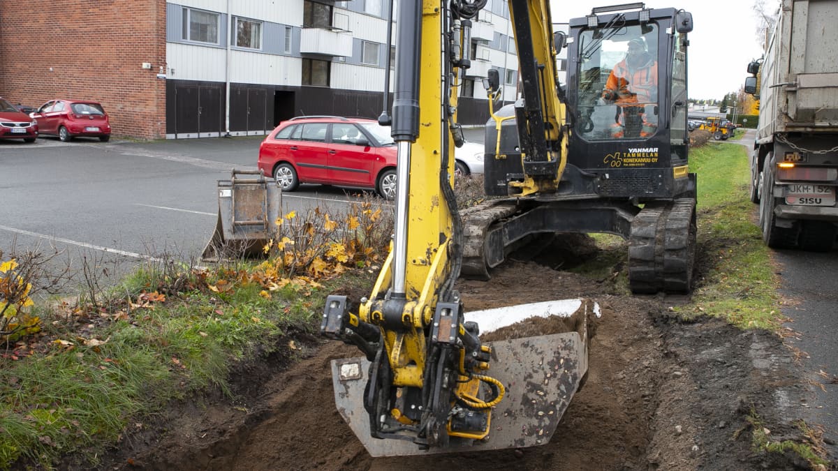 Kaivinkone kaivaa maata kerrostalon parkkipaikan vieressä.
