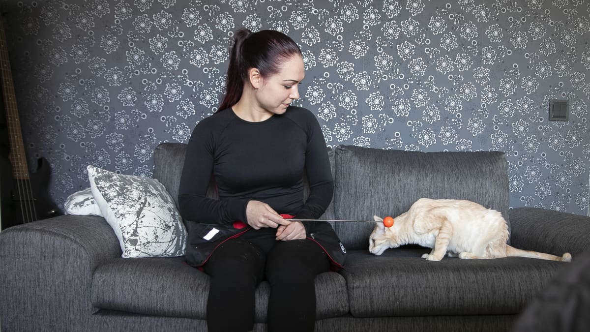 Tummiin pukeutunut, ponihäntäinen nainen istuu sohvalla ja katsoo vieressään olevaa kissaa. Kädessä on kosketuskeppi, jota käytetään eläinten kouluttamisessa. 