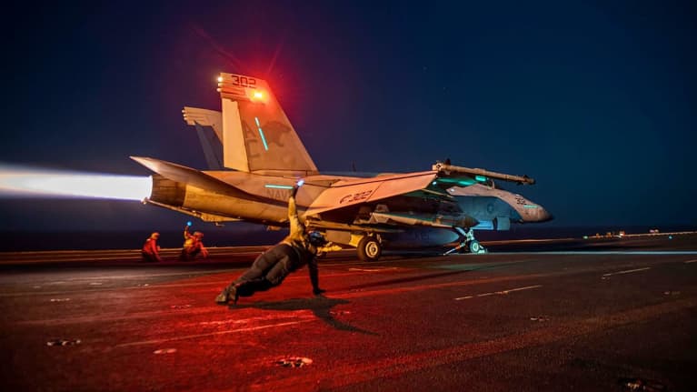 Yhdysvaltain asevoimien hävittäjäkone valmistautumassa nousuun yön pimeydessä, etualalla lentotukialuksen henkilökuntaa.