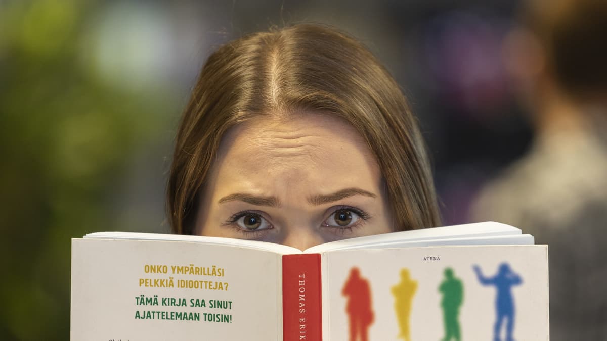 Nainen katsoo kameraan Idiootit ympärilläni -kirjan takaa tuskastunut ilme kasvoillaan. Takana on himmeällä kaksi muuta hahmoa.