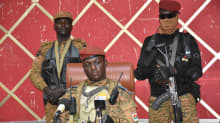 Yksi sotilas istuu pöydän ääressä pitämässä lehdistötilaisuutta ja kaksi sotilasta seisoo hänen takanaan.