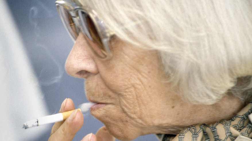 Valkohiuksihen iäkäs henkilö lähikuvassa kuvattuna sivusta, suuret aurinkolasit silmillä, tupakka sormissa, huulten välistä tulee savua.