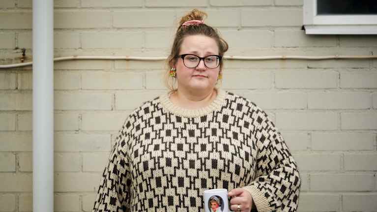 En person i mönstrad tröja håller en mugg framför en ljus tegelvägg.