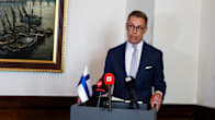A man in a dark blue suit stands speaking behind a podium with a Finnish flag and several microphones, with a maritime painting on the wall.
