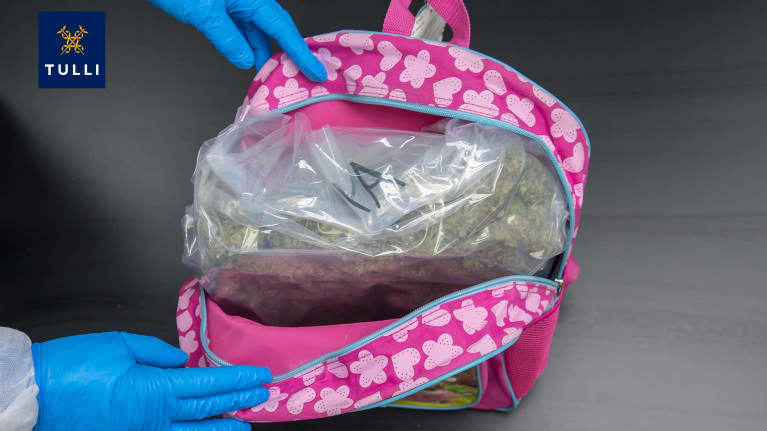 Avattu lasten pinkki reppu, jossa sisällä läpinäkyvä, huumeilla täytetty muovipussi. Kuvassa näkyy kädet, joissa siniset kumihanskat.