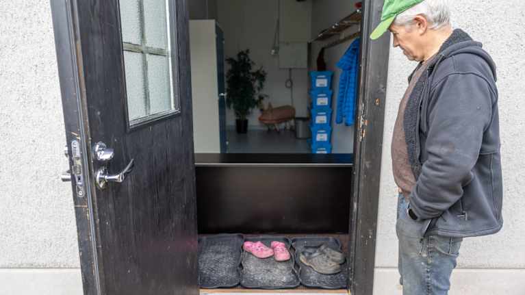 En man tittar in i en tambur. På golvet framför ytterdörren finns gummimattor med skor på, bakom dem en svart bänk man måste stiga över för att komma in i rummet.