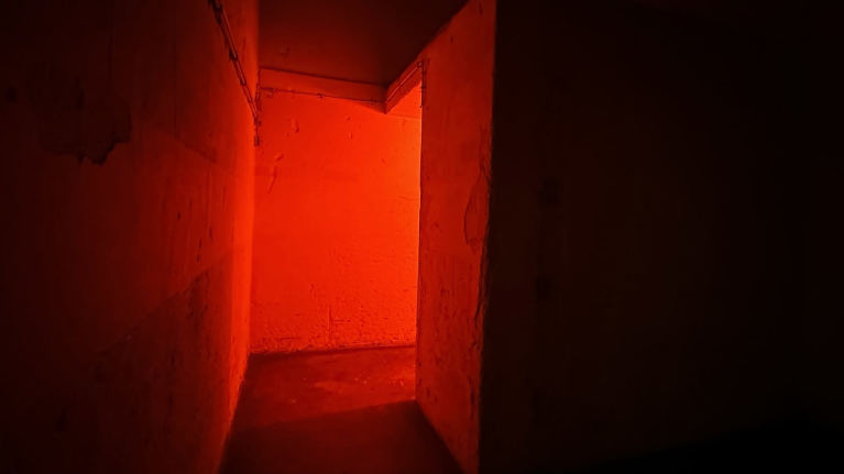 Dramaattista punaista valoa bunkkerissa, sementtinen käytävä kääntyy oikealle valoa kohti. Ympärillä pimeää.