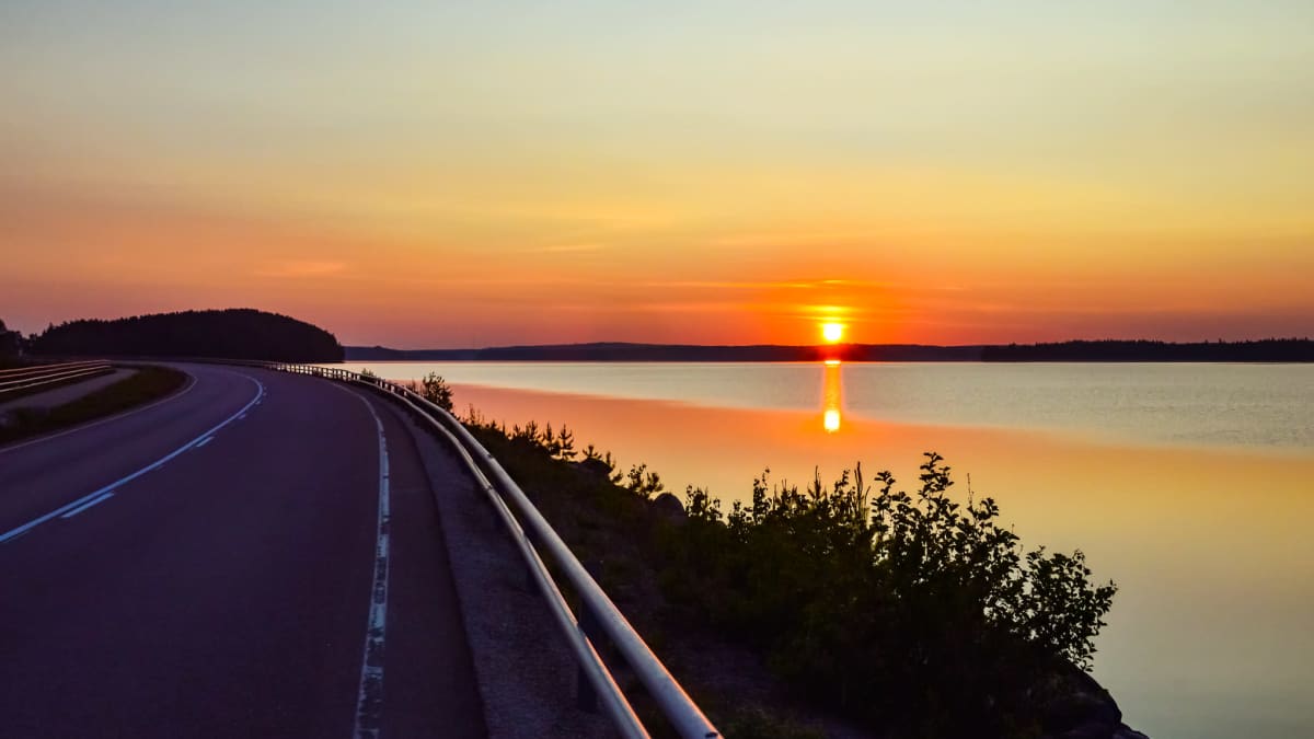 Aamu Asikkalassa. Aurinko nousee järven takaa, asfalttitie kaartaa vasemmalle. 