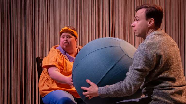 Två dansare sitter på stolar mitt emot varandra och håller en stor boll mellan sig. 