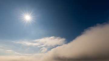 Usvainen merimaisema kirkkaassa auringonvalossa, jossa aurinko paistaa siniseltä taivaalta ja valkoinen pilviverho leviää horisontissa veden yllä.