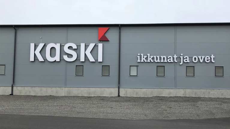 Harmaan teollisuushallin seinässä on teksti Kaski, ikkunat ja ovet ja Kaski-sanan i-kirjaimen päällä on punainen viirilippua muistuttava kuvio.