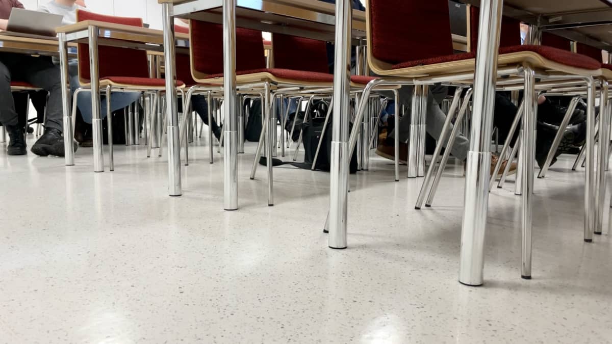 Luentosalin tuoleja ja pöytiä sekä niiden seassa opiskelijoiden jalkoja alhaalta kuvattuna.