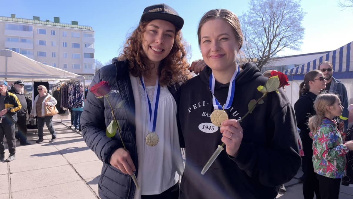 Kaksi hymyilevää naista, joilla kultamitalit kaulassa ja ruusut kädessä. 