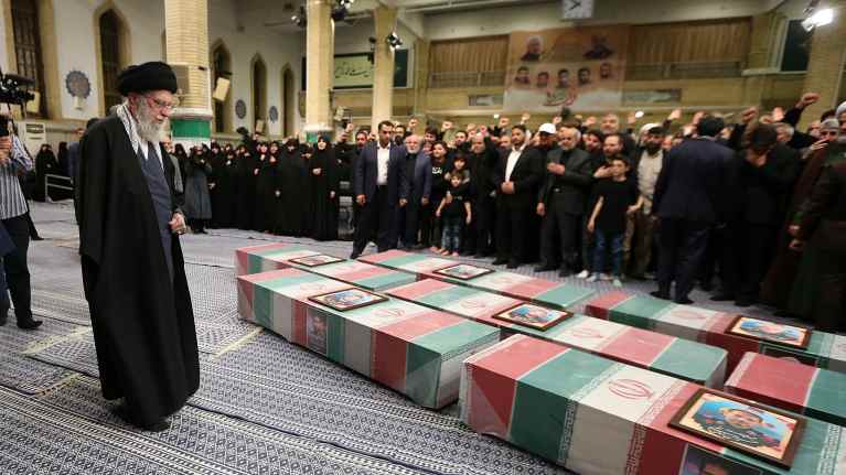 Ayatollah Ali Khamenei katsoo maassa makaavia arkkuja, jotka on päällystetty Iranin lipuilla. Arkkujen päällä on valokuvat kuolleista. Taustalla miehet mustissa puvuissa seisovat omana ryhmäna ja mustiin hijabeihin pukeutuneet naiset omana ryhmänä.