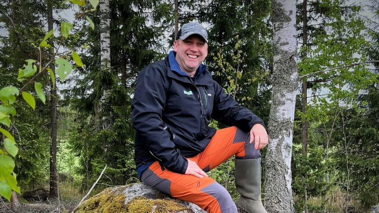En man sitter på en sten i skogen och ler.