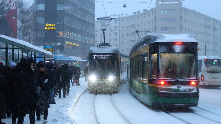 Helsinki street, covered in snow during a blizzard, with pedestrians, trams and cars on the road.