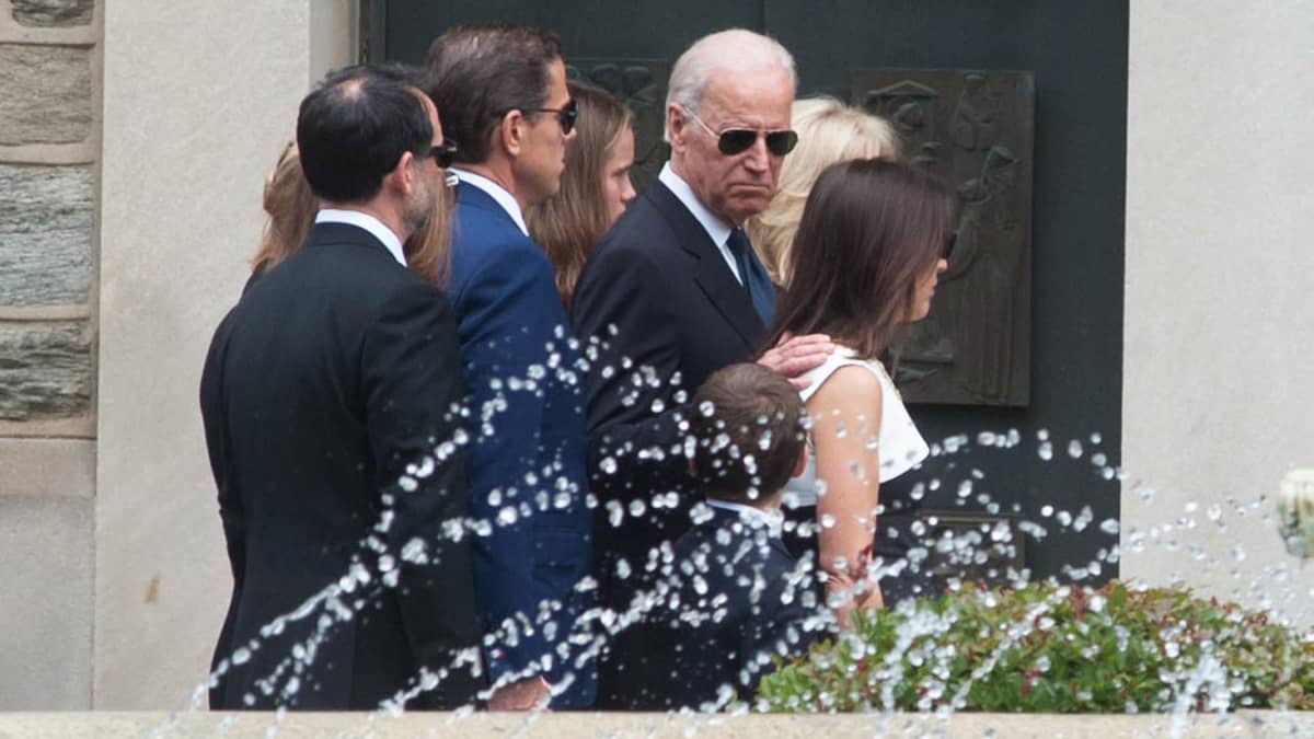 Joe Biden perheineen Bidenin esikoispoika Beaun hautajaisissa. Kuvan etualalla on suihkulähde. Biden perheineen on kuvassa tiiviisti yhdessä. Bidenillä itsellään on mustat aurinkolasit ja hän on ainoa joka katsoo kuvaajaa kohden, ilmeensä vakavana.