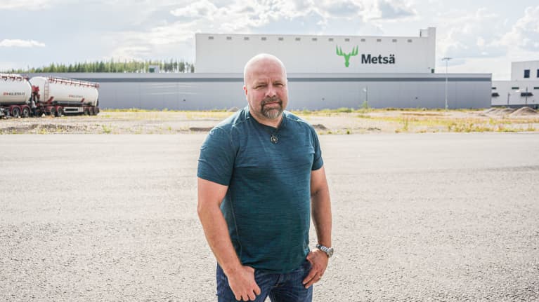 Kalju, parrakas keski-ikäinen mies vihreässä t-paidassa katsoo kameraan suuren metsäteollisuuden tehdasrakennuksen edessä.