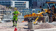 A construction worker stands on a site in front of a road grader. 