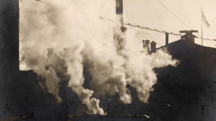 Vanhassa mustavalkoisessa valokuvassa talojen katolla olevista savupiipuista tupruaa sankkaa savua.