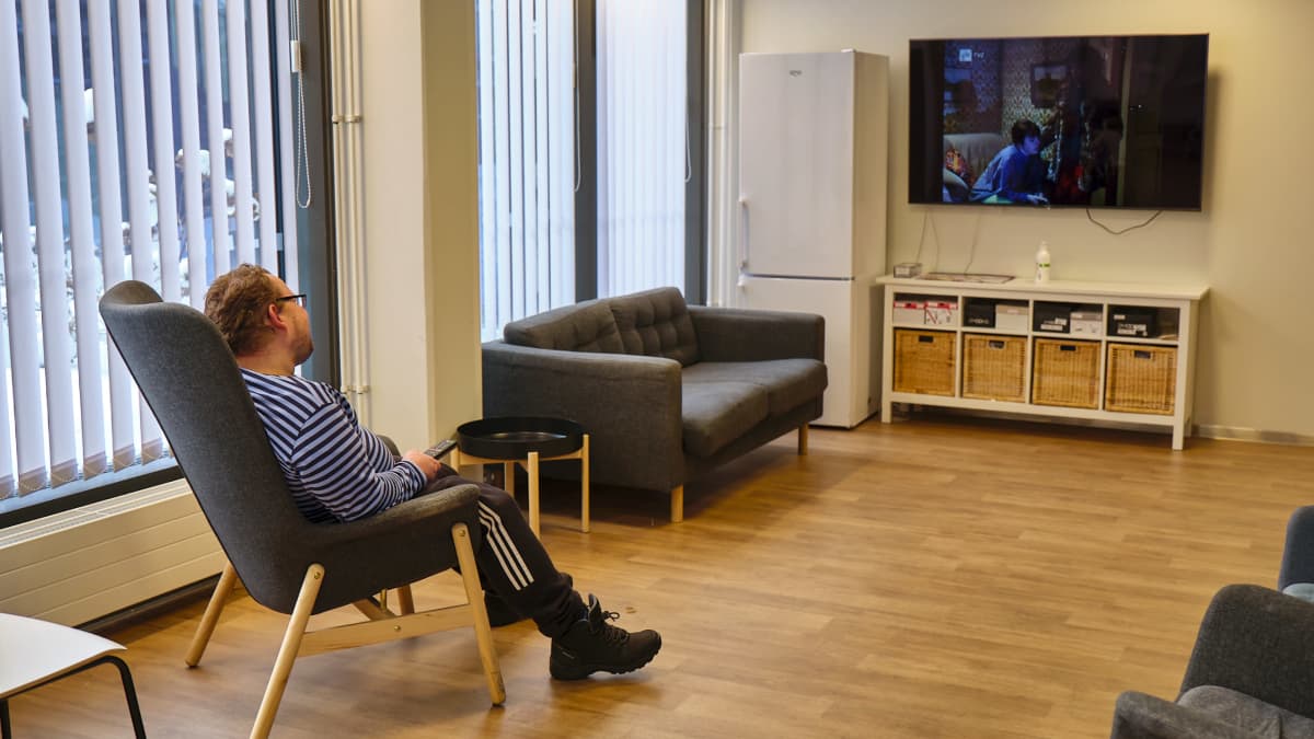 Lasse Purmonen katsoo televisiota sinimustaraidallisessa paidassa. Taustalla näkyy laajakuvatelkkari ja harmaa sohva.
