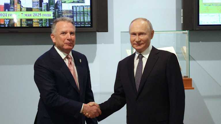 Steve Witkoff och Vladimir Putin skakar hand. Båda ler och tittar direkt in i kameran. Bakom dem ett par vitriner med datum och kartor. 