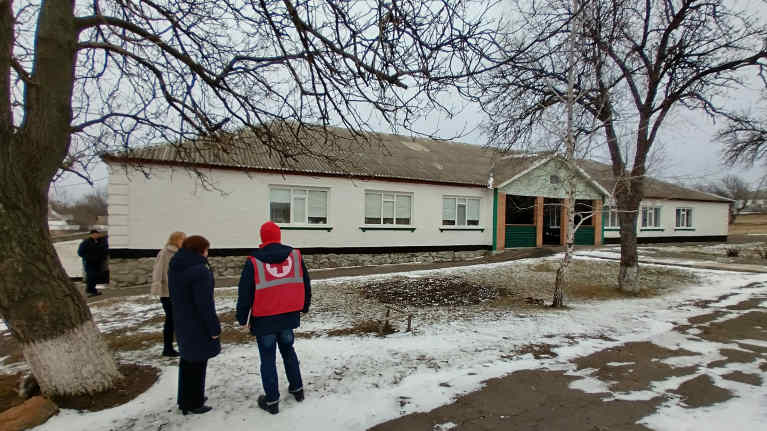 Valkoinen, yksikerroksinen toimistorakennus. Edessä kuvaan selin seisoo kolme ihmistä, joista yhdellä on Punaisen Ristin huomioliivi. 