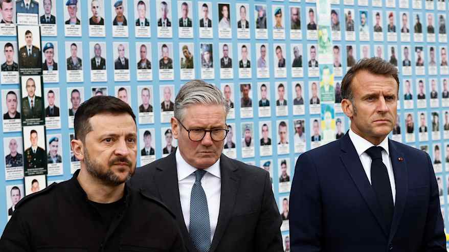 Emmanuel Macron, Keir Starmer, ja Voloydmyr Zelenskyi kaatuneiden ukralaissotilaiden kuvia esittävän seinän edustalla. 