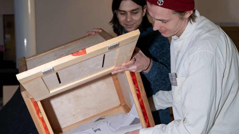 Two people tip over a wooden ballot box, pouring out ballots onto a table.