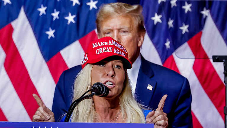 Marjorie Taylor Greene står och håller tal i Magahatt framför en amerikansk flagga. Bakom syns Donald Trump. 