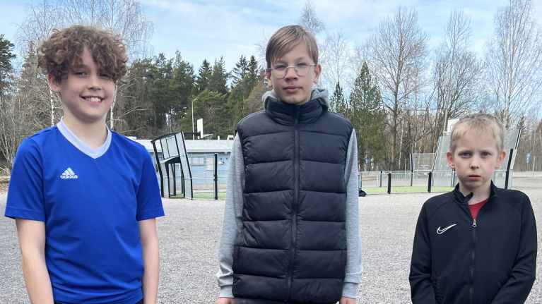 Kolme poikaa seisoo koulun sorakentällä.