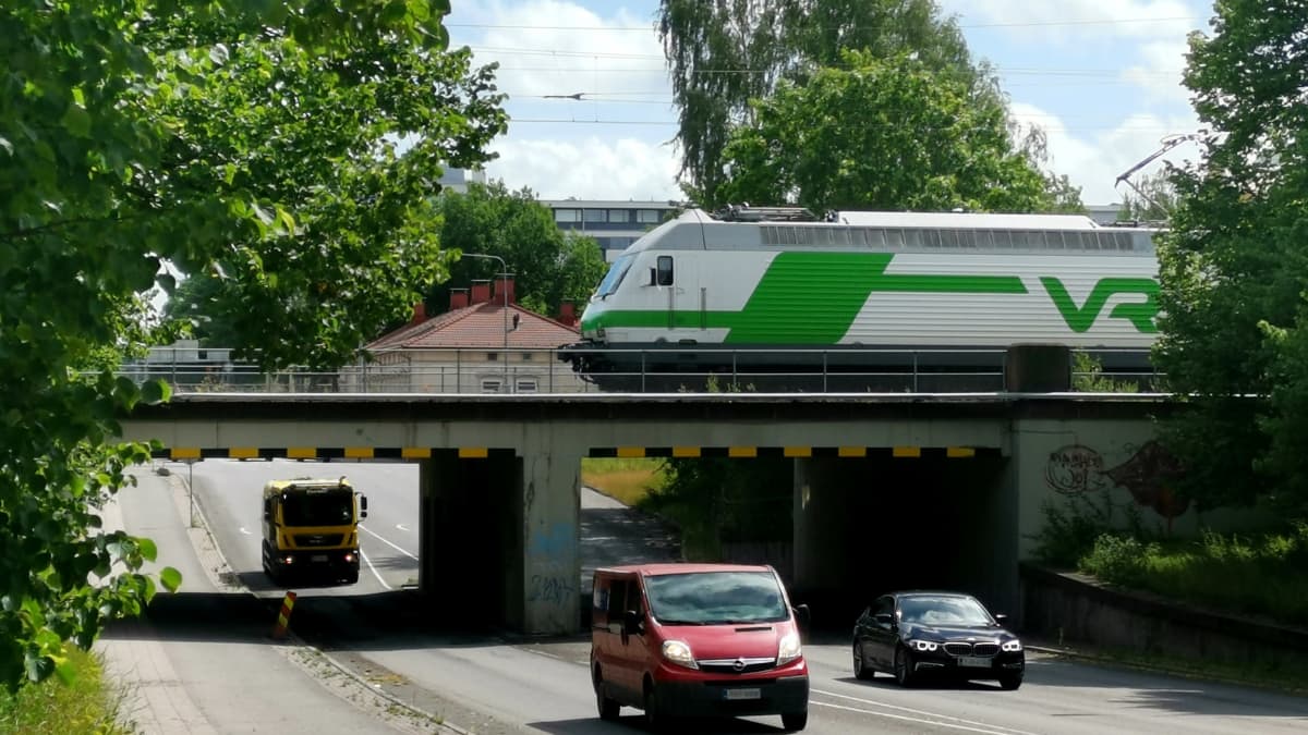 VR:n veturi ylittämässä Koulukadun alikulkusiltaa Turussa, alla autoja, ympärillä vehreitä puita ja sinistä taivasta ja poutapilviä.