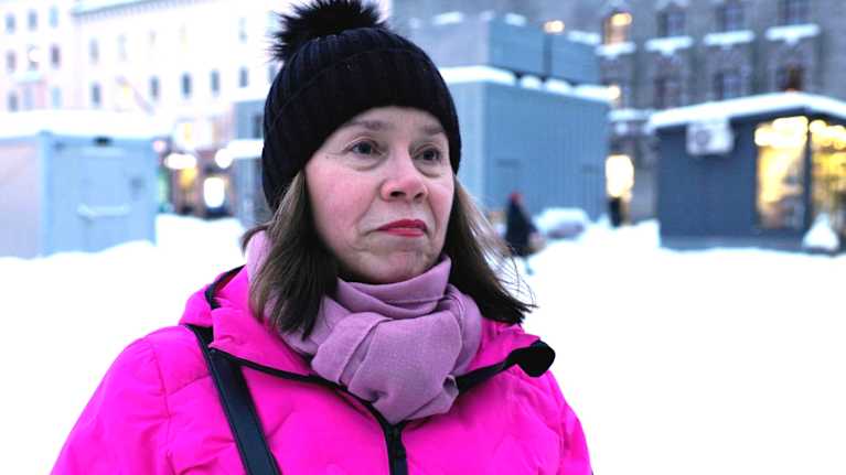 Mustapipoinen, pinkkiin takkiin pukeutunut nainen (Riikka Hermunen). Taustalla luminen toriympäristö.