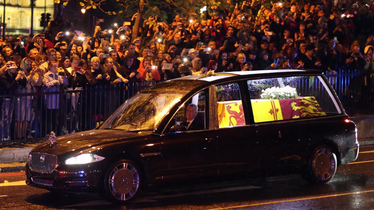En svart likbil med drottningens kista kör på en gata som blänker av regn i mörkret. Kistan i bilen är upplyst och man kan se vita blommor i en krans på kistan som är täckt med den kungliga flaggan. I bakgrunden syns en folkmassa vid gatkanten.