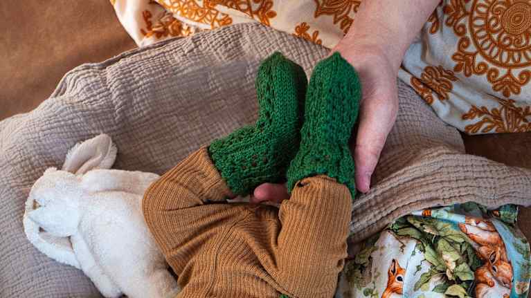 An adult's hand holding an infant's legs and feet, which are clothed in brown pants and green woolen socks.