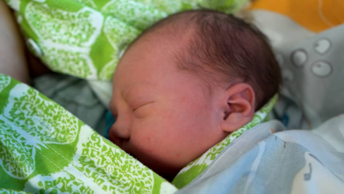 Nukkuvan vauvan kasvot sivusta kuvattuna. Tummahiuksisen vauvan päällä vaaleaa peittoa kaulaan asti ja ympärillä vihreävalkoisia liinavaatteita. 