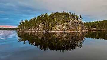 Tyyni järvimaisema iltahämärässä: kuvassa näkyy metsäinen saari, joka heijastuu peilikirkkaasta vedestä.