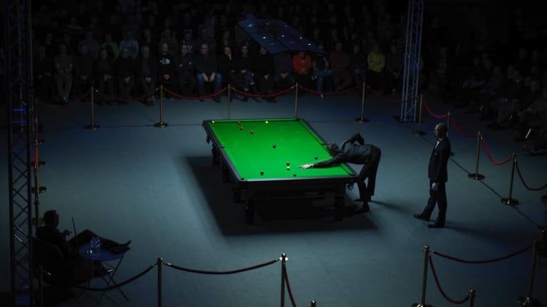 Keskellä on vihreä Snooker-pöytä, joka on valaistu kirkkaalla valolla. Pöydän äärellä on pelaaja, joka keskittyy tähtäämään palloon.