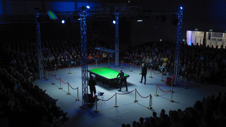 Keskellä on vihreä Snooker-pöytä, joka on valaistu kirkkaalla valolla. Ympärillä näkyy paljon yleisöä istumassa.