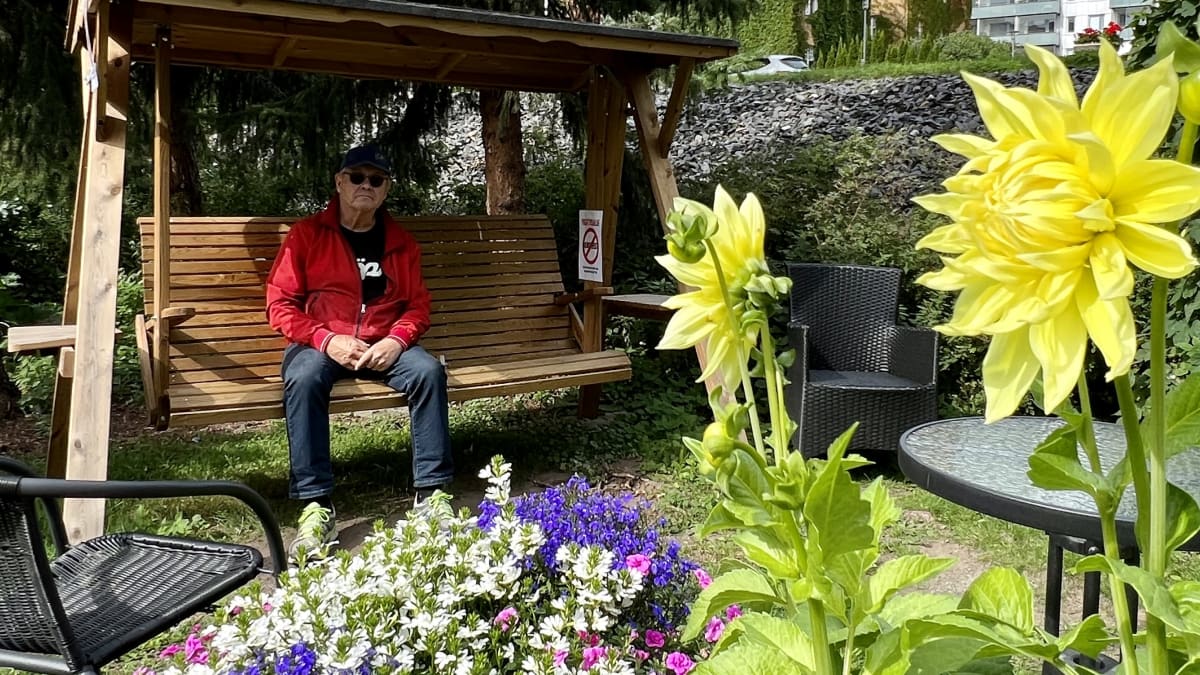 Mies istuu puisessa puutarhakeinussa. Keinuun on kiinnitetty kyltti, jonka teksti kertoo talon piha-alueen olevan varattu vain talon oman väen käyttöön. Pihalla on kauniita kukkaistutuksia.