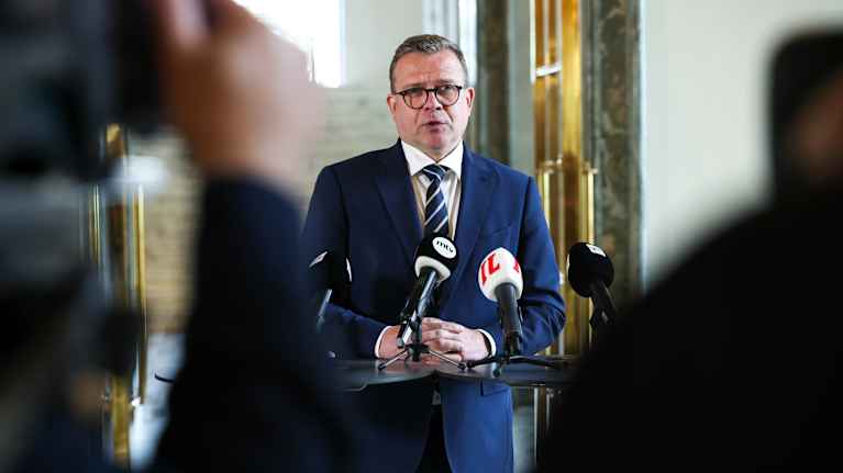 A man with a blue suit and a concerned expression stands behind a podium with microphones.