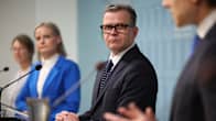Statsminister Petteri Orpo fotograferad snett från sidan på en presskonferens med ministrarna Riikka Purra och Sari Essayah bredvid sig så att de blir i bakgrunden på bilden.
