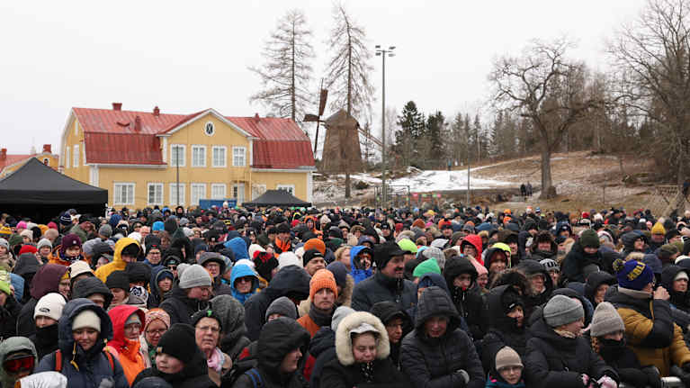 A tightly packed crowd of people bundled up in winter clothing in an outdoor area with a yellow and red wooden house behind them and some snow in a grassy slope. 